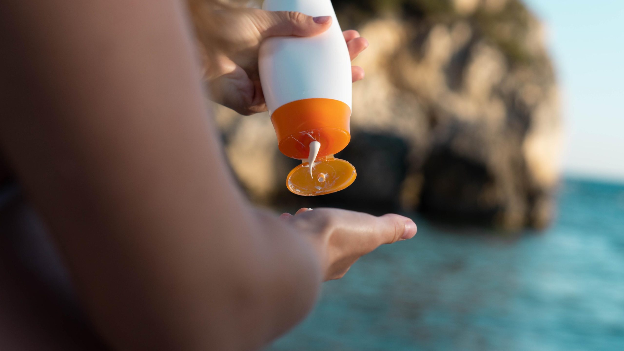 Woman's hand holding a bottle of sunscreen with the sea at the background.