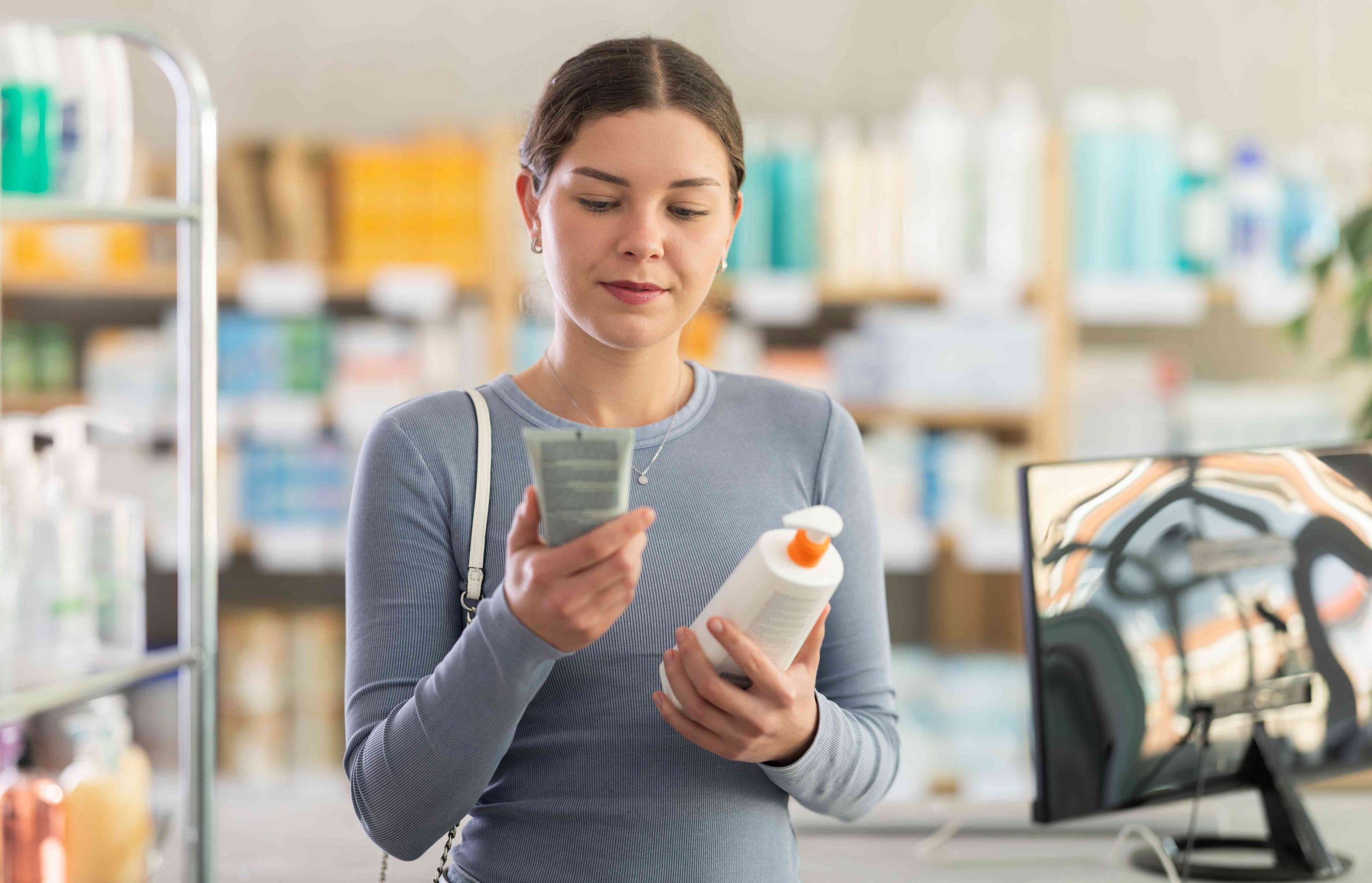 Young woman comparing two skincare products with interest at pharmacy, pump bottle and tube, exploring difference in texture, purpose or ingredients before making purchase.