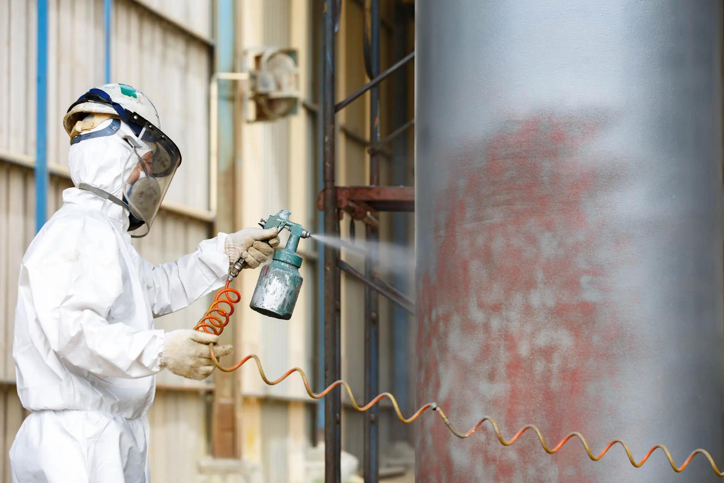 Industrial worker applying an anti-corrosion protective coating on a large steel structure using a spray system in an industrial environment