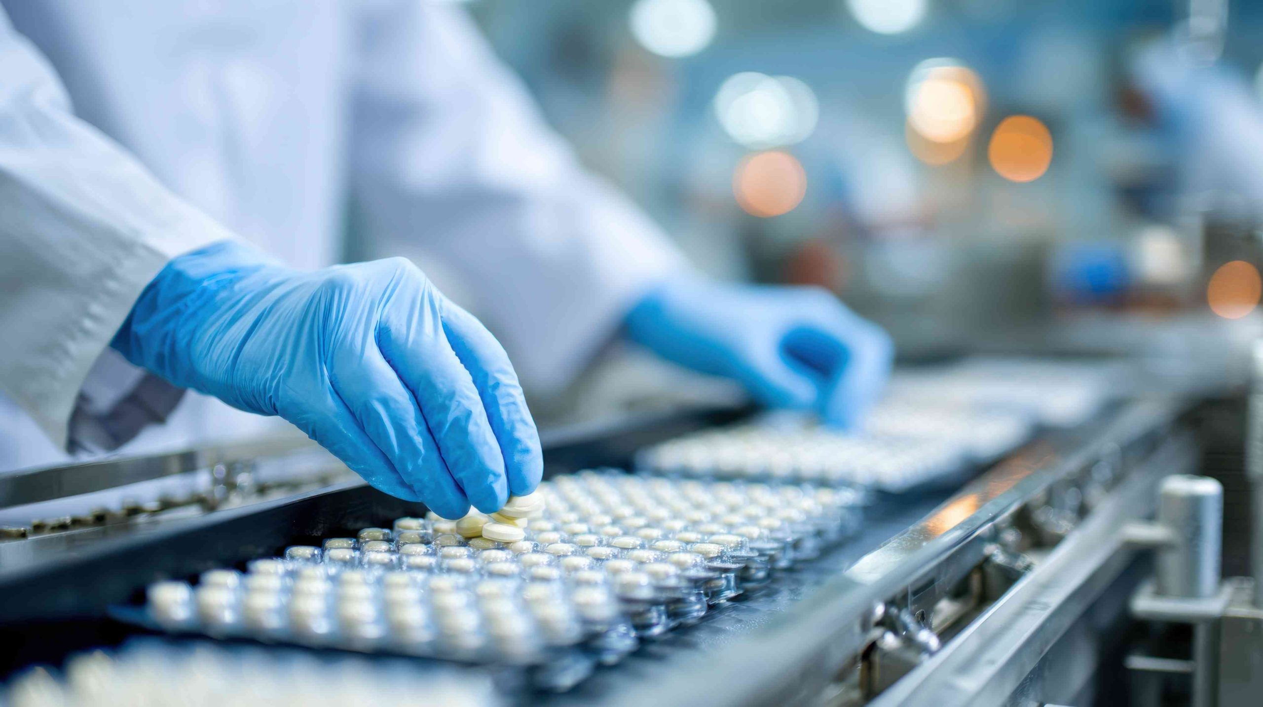 A laboratory worker wearing gloves carefully arranges vials on a conveyor belt in a pharmaceutical facility.